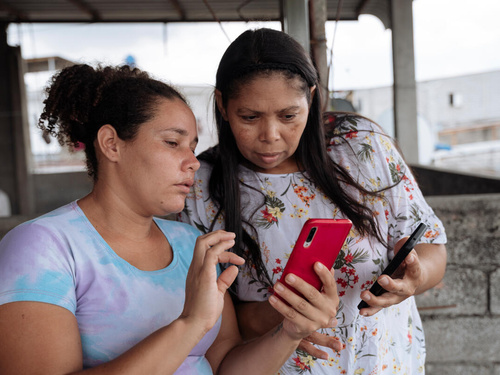 Dos mujeres. Una de ellas sostienen un teléfono móvil y se lo muestra a la otra.