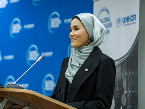 Una mujer, que lleva puesto un hijab, pronuncia un discurso desde un podio. Frente a ella, un delgado micrófono; detrás, un fondo azul con el logotipo del Foro Mundial sobre los Refugiados.,