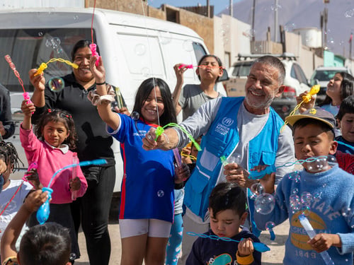 Un hombre con chaleco azul y camiseta blanca debajo, juega con burbujas de jabón junto a un grupo de niños
