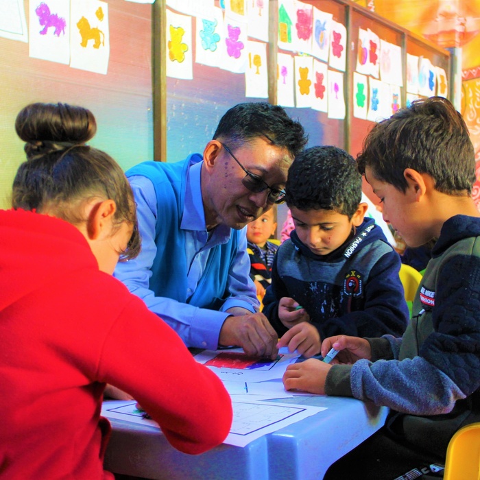 El Sr. Wanchai Tachavejnukul dibujando junto a niñas y niños, sentado a una mesa en en aula escolar.