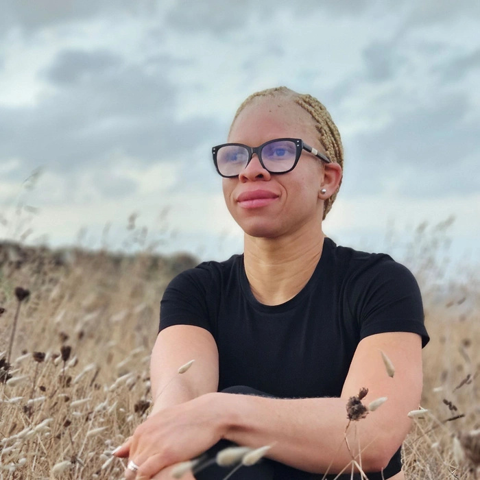 Una mujer albina con gafas está sentada en un campo, mirando a lo lejos, con el pelo recogido.