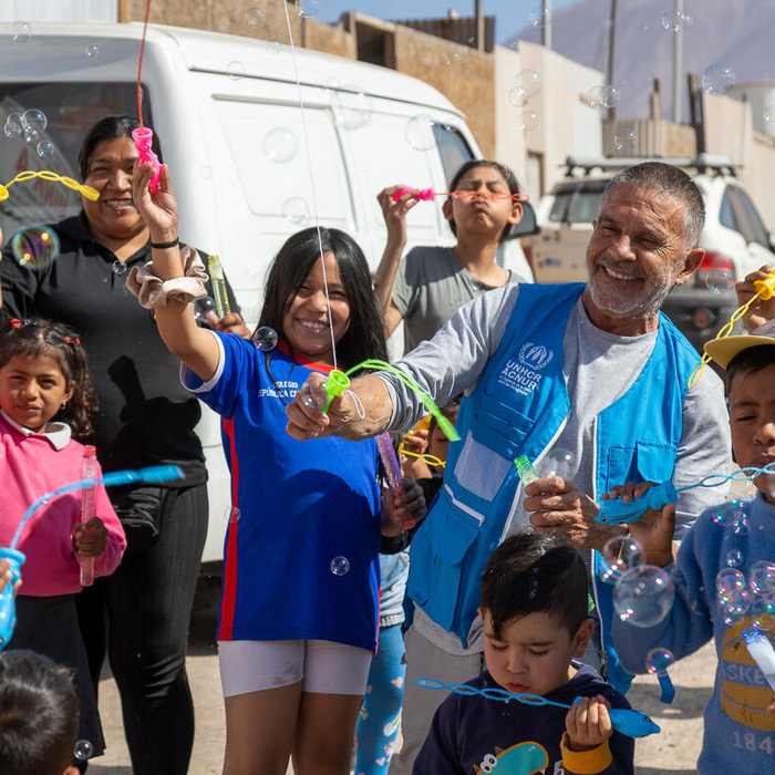 Un hombre con chaleco azul y camiseta blanca debajo, juega con burbujas de jabón junto a un grupo de niños