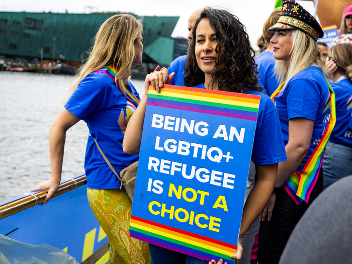 En una embarcación que navega por un río, una joven muestra un cartel que se lee: "Nadie opta por ser una persona LGBTIQ+ refugiada"