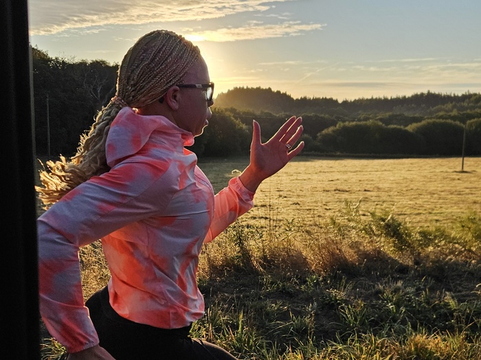Una mujer con largas trenzas recogidas corre en la campiña, con el sol a sus espaldas.