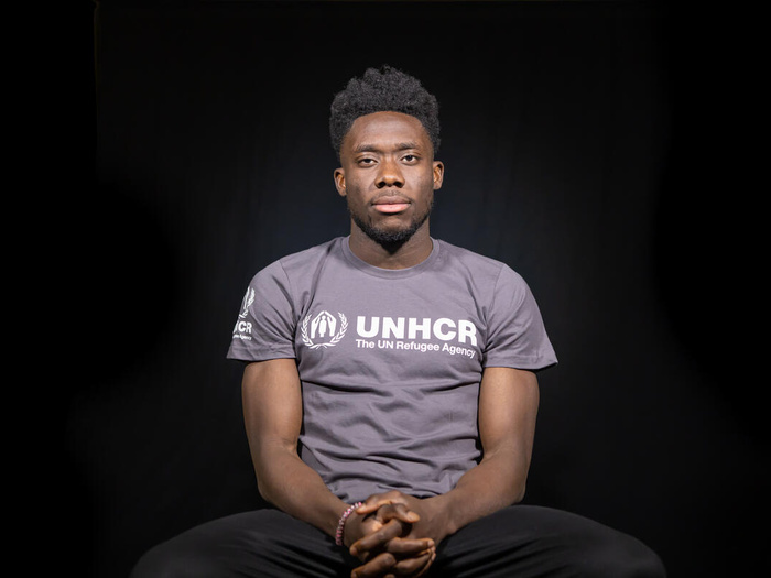 Un hombre afro que lleva puesta una camisa color gris oscuro con el logotipo de ACNUR en color blanco mira fijamente a la cámara desde la silla en la que está sentado