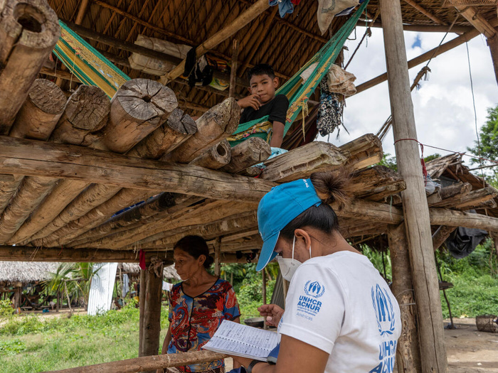 Una trabajadora de ACNUR trabaja en un documento; delante de ella, una mujer warao está bajo la sombra de una estructura de madera. En la parte superior de la estructura, un niño decansa en una hamaca.