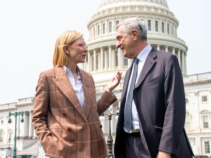Una mujer rubia charla con un hombre cano; ambos llevan puesto un traje sastre. Detrás suyo, el Capitolio de Estados Unidos.