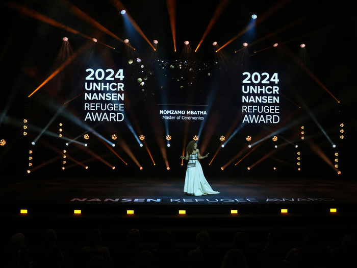 Una mujer negra camina, micrófono en mano, sobre un escenario repleto de luces color naranja. Al fondo se está proyectando, en color blanco, un texto que reza: "Premio Nansen de ACNUR para los Refugiados de 2024".