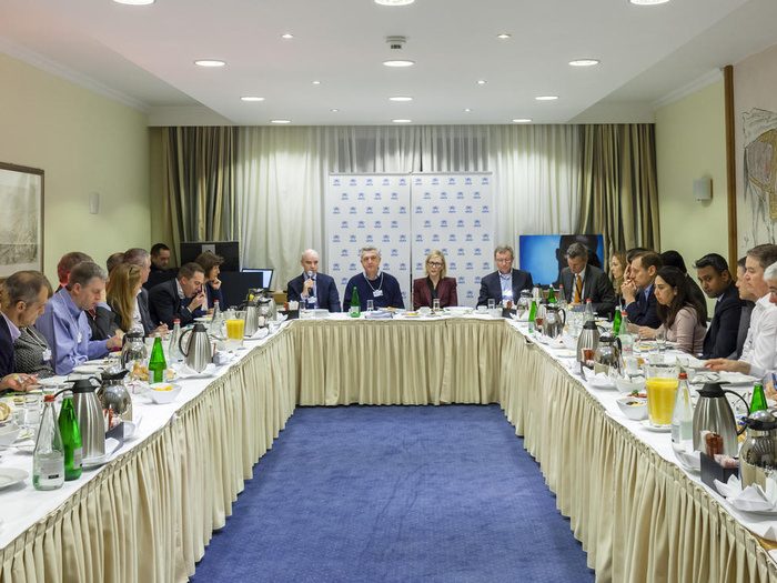 Un numeroso grupo de personas dialoga en un salón con mesas colocadas en forma de herradura