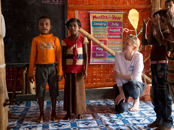 Hincada, una mujer rubia observa con cariño a un chico y una chica rohingya