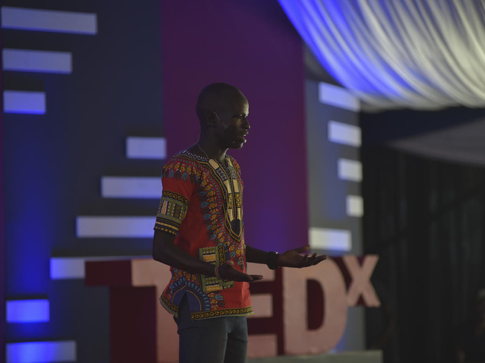 Un hombre sursudanés, que lleva puesta una colorida indumentaria, da una charla desde un escenario; detrás suyo, las letras TEDx.