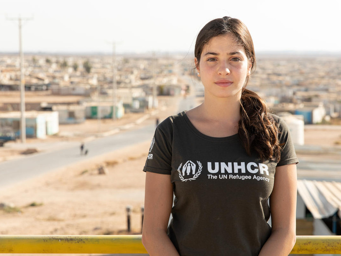 Una joven que lleva el cabello recogido y porta una camiseta negra con el logotipo de ACNUR en color blanco mira fijamente a la cámara; detrás suyo, un campamento de refugiados.