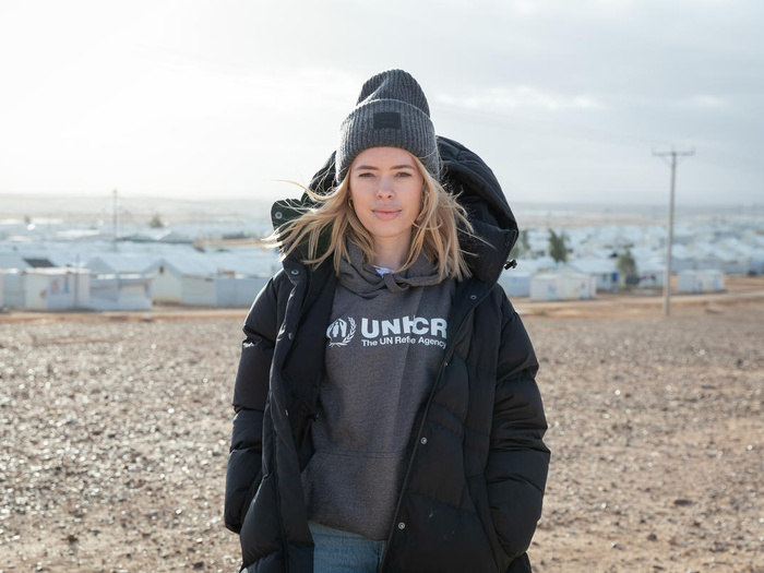 Una mujer caucásica, que lleva puesto un gorro de estambre y una sudadera color gris con el logotipo de ACNUR, sonríe a la cámara; detrás suyo, un campamento de refugiados.