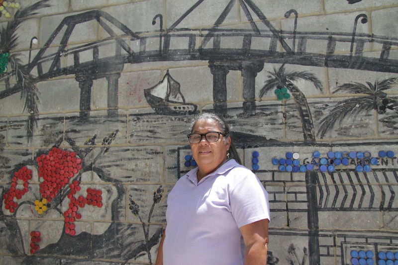 Una mujer con una camisa tipo polo color lila, frente a un mural de un puente que pasa por encima del mar.