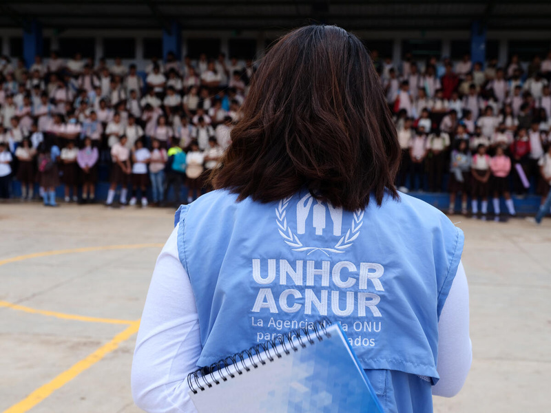 Una mujer que porta chaleco azul con logo de ACNUR está de espaldas frente a una multitud y sostiene un cuaderno en sus manos