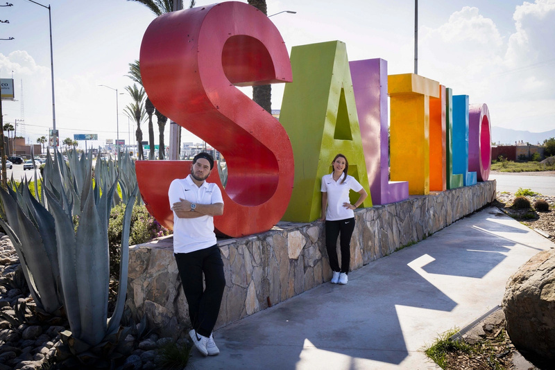Un hombre y una mujer que visten playera blanca y pantalón negro, posan de pie frente a un letrero grande que dice "Saltillo"