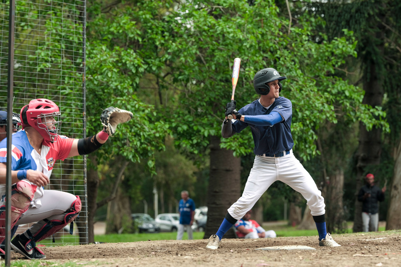Un hombre de uniforme azul marino con blanco toma un bate en sus manos, a punto de golpear una pelota de softbol