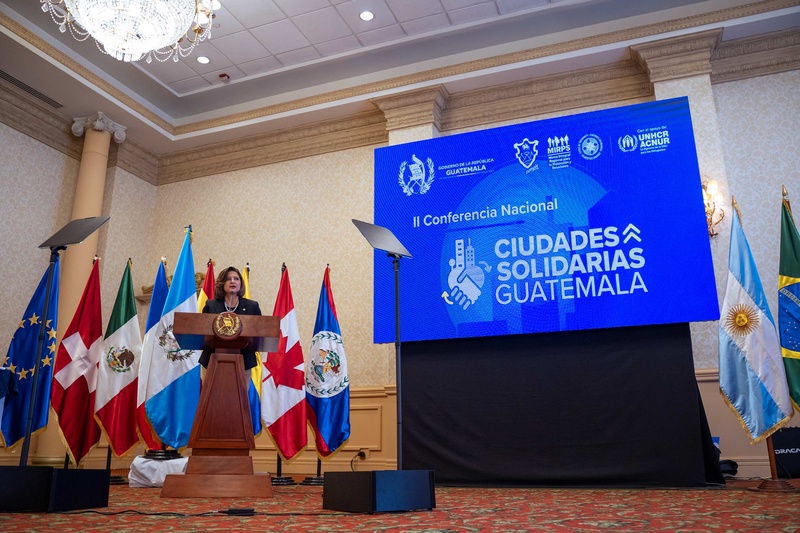 Una mujer con vestimenta formal, habla en un podio con 9 banderas de diferentes países detrás de ella. Al lado se lee en un fondo azul con letras blancas: CIUDADES SOLIDARIAS GUATEMALA