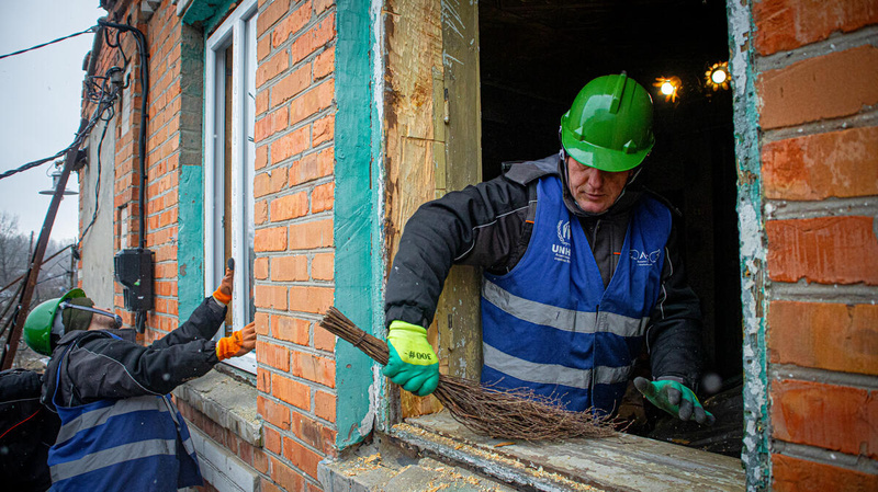 Dos hombres que llevan puestos cascos de construcción trabajan en la reparación de las ventanas de un vivienda hecha de ladrillo.