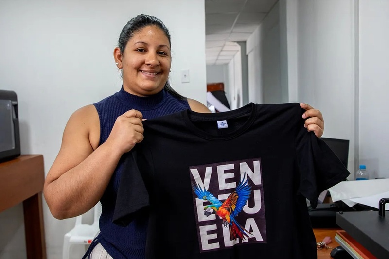 Una mujer muestra una camiseta con una impresión original donde se lee 'Venezuela', decorada con la imagen de una guacamaya..