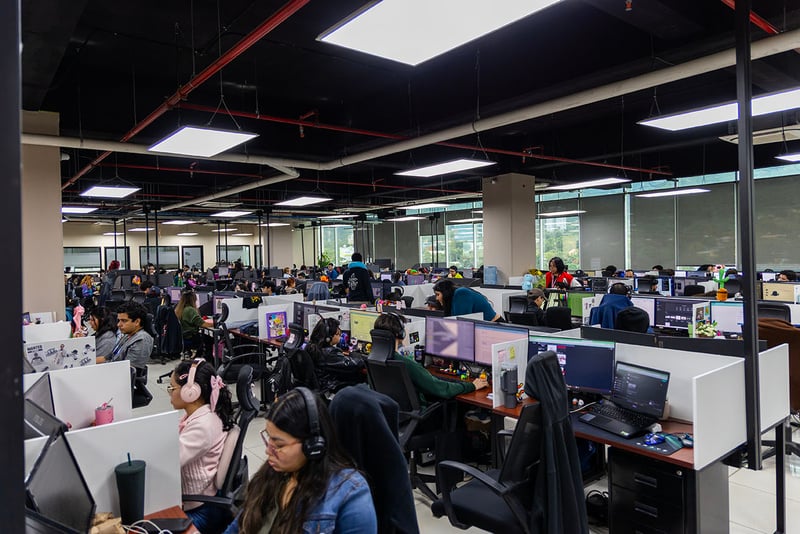 Una oficina abierta con varios escritorios ordenados en filas donde muchas trabajan frente a pantallas de computadoras.