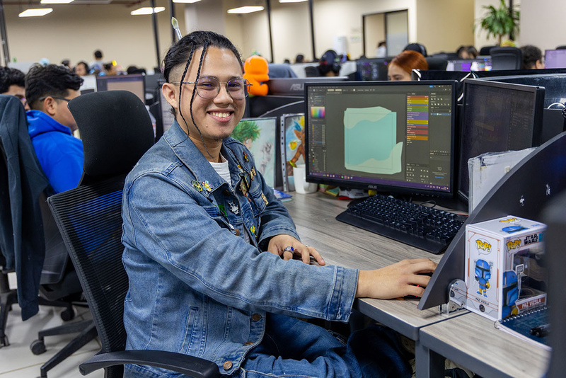 Un joven posa para la cámara sentado a su escritorio, frente a dos pantallas de computadora.
