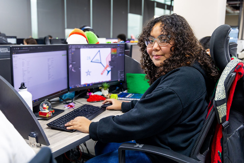 Una joven posa para la cámara sentada a su escritorio, donde se encuentran dos pantallas de computadora y un teclado.