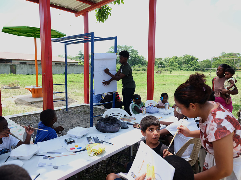 Una joven ayuda a un grupo de niños y niñas a pintar y dibujar en una actividad al aire libre.