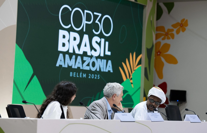 Tres personas sentadas en el palco de oradores de un evento. Detrás de ellas, un letrero de la COP30.
