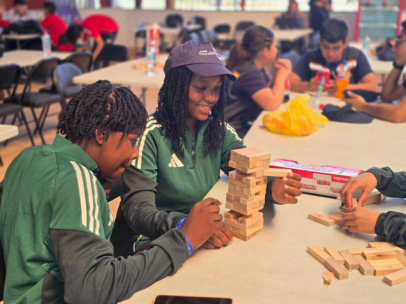 Un joven y una joven con ropa deportiva verde juegan jenga en un aula donde hay más jóvenes