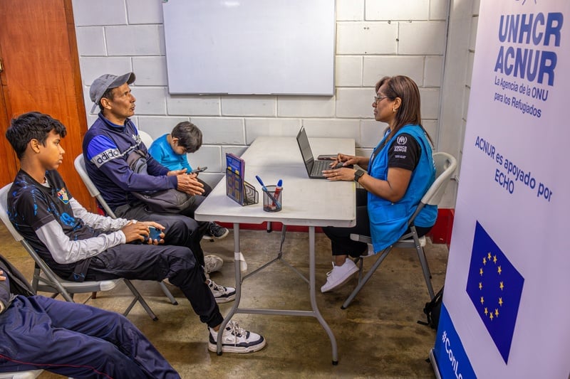 Un adulto y tres niños hablan con una funcionaria de ACNUR, reconocible por el chaleco azul que lleva puesto, que está tomando notas en una computadora
