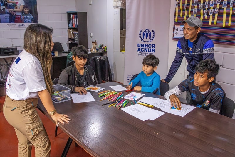 Una mujer habla con un adulto y tres niños. Los niños están sentados a una mesa, donde están dibujando, el adulto está de pie detrás de ellos