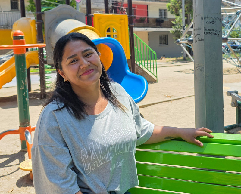 Una mujer de cabello negro que viste playera gris, posa sentada en una banca verde de un parque, al fondo se ven juegos infantiles