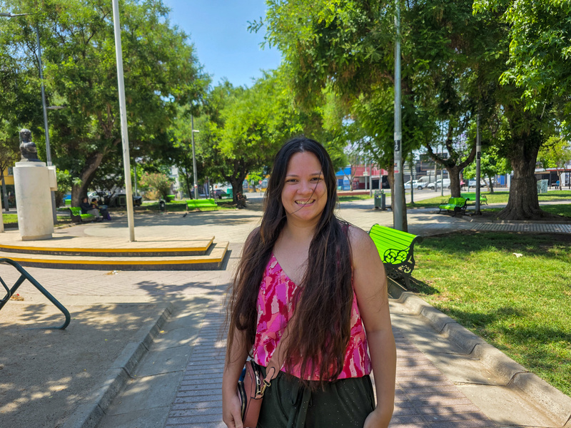Una mujer joven de cabello largo que viste blusa rosa, de pie en un parque donde se observan árboles al fondo