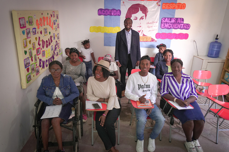 De pie al centro un hombre con vestimenta formal, sentados en pupitres, 9 jóvenes en una clase. Al fondo, una niña juega de pie recargada en una pared
