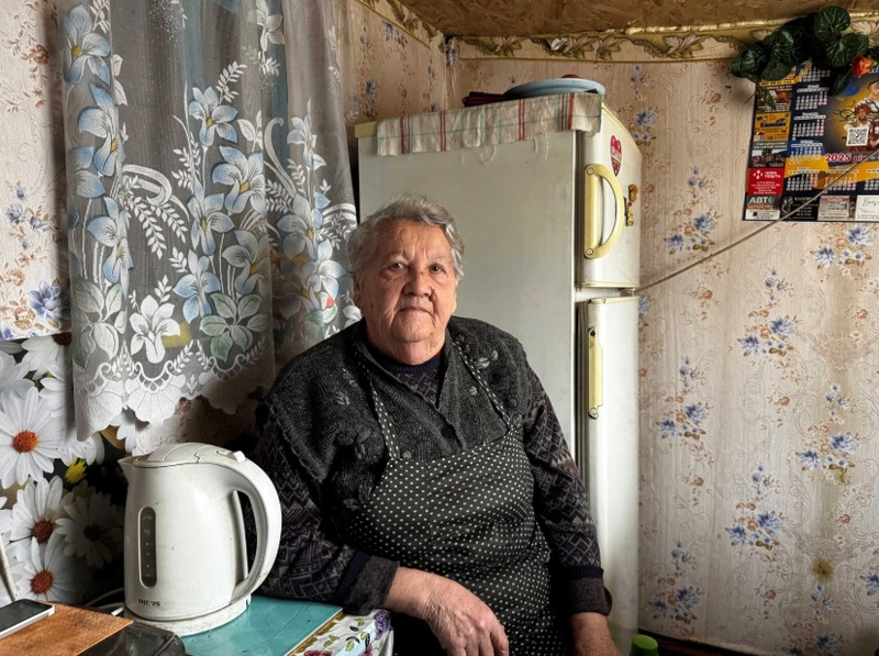Una mujer mayor de cabello corto que viste ropa negra y porta un delantal negro, sentada en su cocina. Detrás de ella unas cortinas floreadas y un refrierador blanco. Junto a su mano derecha una jarra eléctrica color blanco
