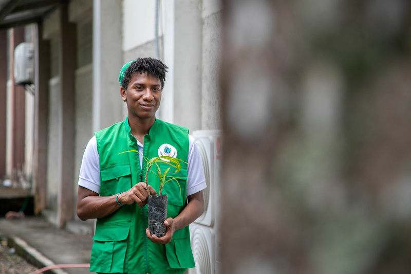 Un joven con un pañuelo verde en la cabeza y chaleco verde toma entre sus manos una planta