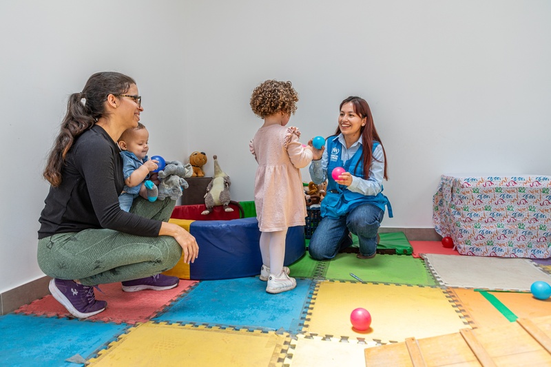 Dos mujeres agachadas en un área de juegos infantiles: una sostiene a un niño pequeño, la otra le muestra un par de pelotas a una niña de cabello rizado rubio que está de pie