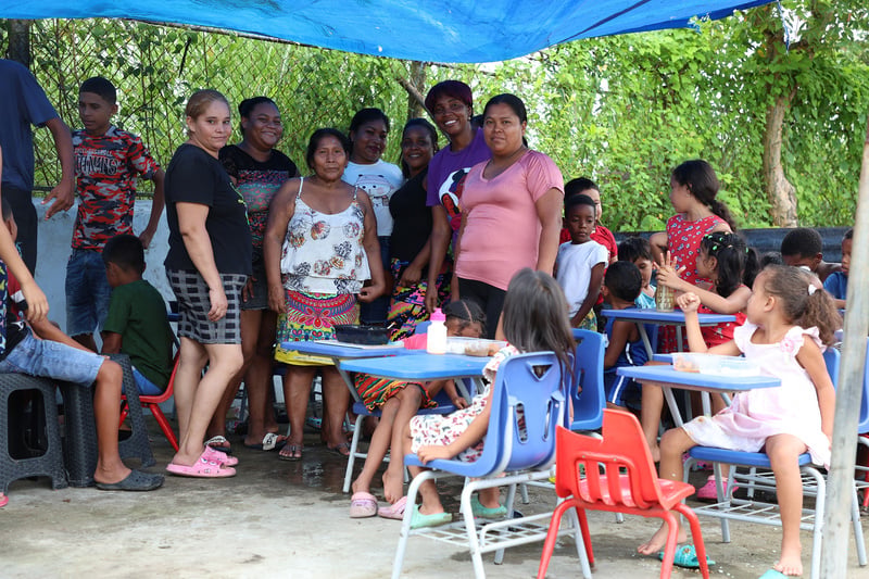 Un grupo de siete madres bajo una carpa azul, junto a niñas y niños que están consumiendo alimentos