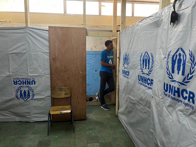 Un hombre vestido con una camiseta azul y pantalones negros se asoma a la puerta de una habitación dividida con marcos de madera y láminas de plástico de ACNUR