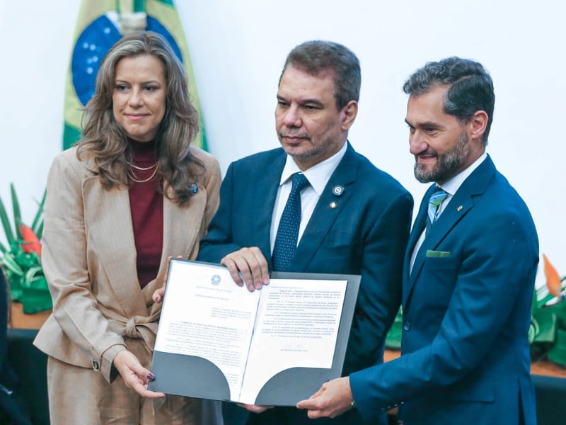 Una mujer y dos hombres muestran un documento en una carpeta. Detrás de ellos la bandera de Brasil.