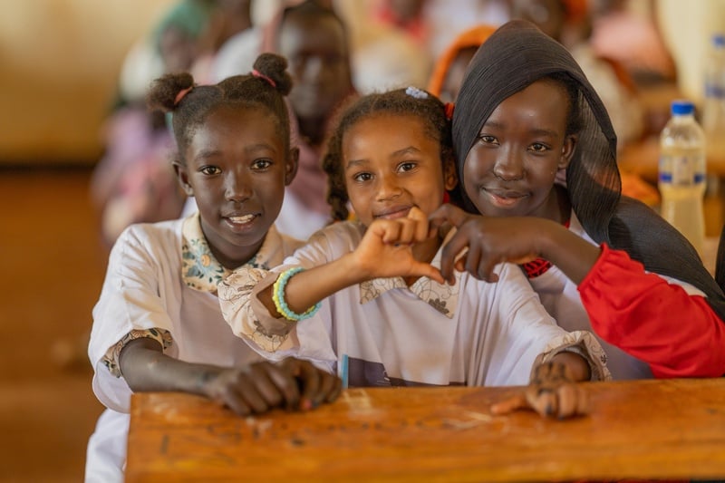 Tres niñas posan para una foto sonriendo en un salón de clases, mientras dos de ellas forman un corazón con sus manos.