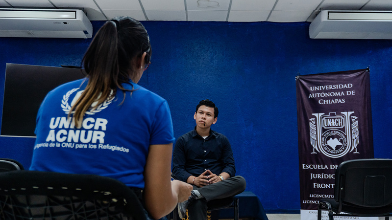 De espaldas, una muejr de playera azul con logo blanco de ACNUR, habla con un joven vestido de negro