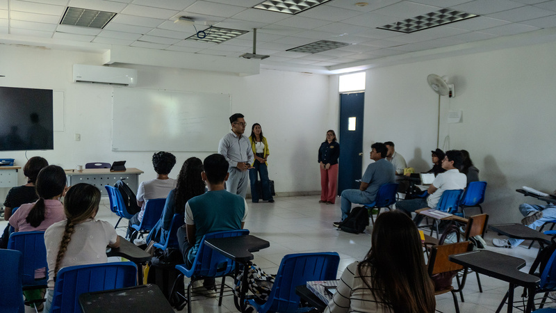 Al menos quince jóvenes sentados en pupitres dentro de un aula, se mantienen atentos a un hombre que habla frente al grupo. Detrás del hombre, hay otras dos mujeres de pie
