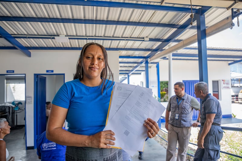 Una mujer que viste camiseta azul, sostiene en sus manos unos documentos