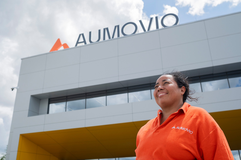 Una mujer que viste una camiseta tipo polo naranja, en una toma en contrapicada donde al fondonse observa un edificio con un letrero que dice AUMOVIO