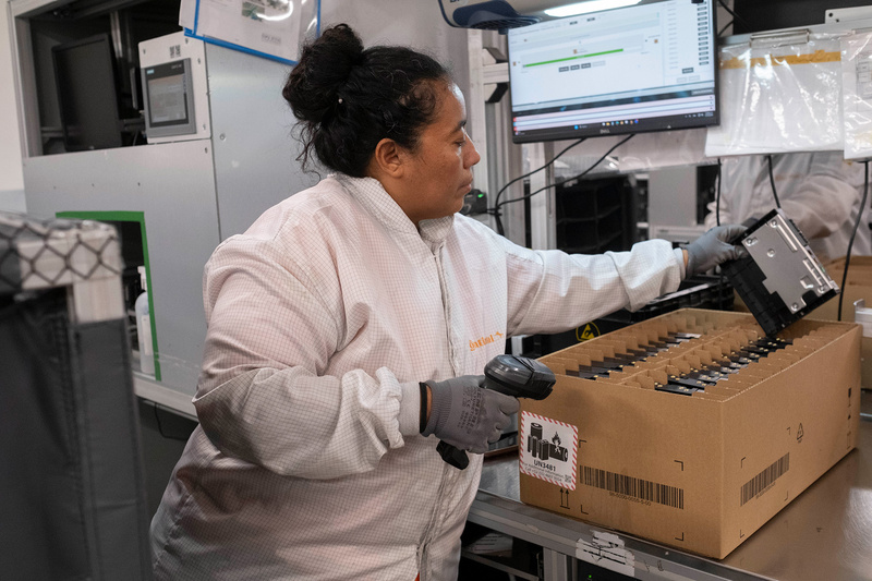 Una mujer vestida con bata blanca de trabajo, guantes y cabello recogido, ordena unos dispositivos en cajas