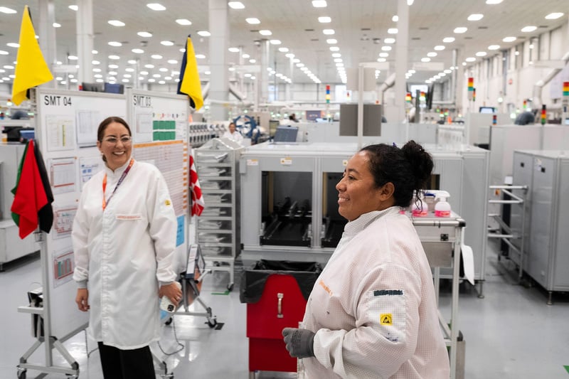 Dos mujeres con batas blancas de trabajo, en el interior de una empresa donde se ve maquinaria