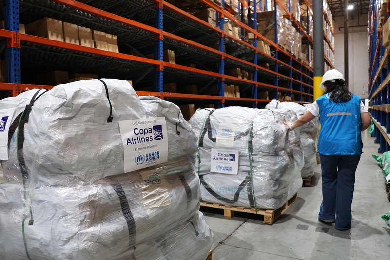 De pie junto a pallets con suministros de emergencia etiquetados con logos de ACNUR y COPA Airlines, una mujer con casco que viste chaleco azul de ACNUR