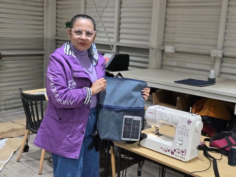 Una mujer que viste vhamarra y blusa moradas, sostiene en sus manos una mochila azul junto a una máquona de coser en un taller de costura